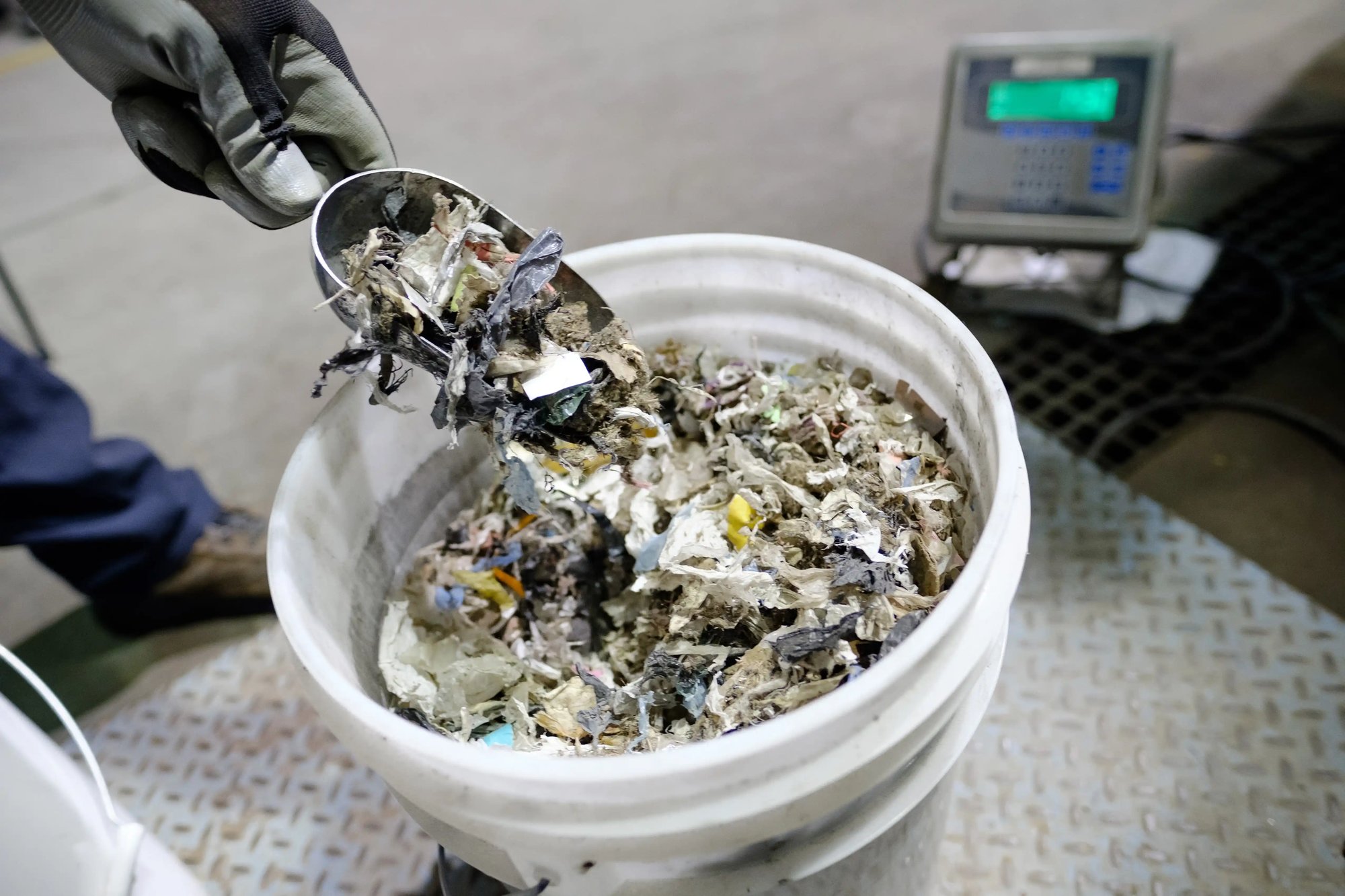 a hand holding a steel hand shovel over a bucket of shredded garbage